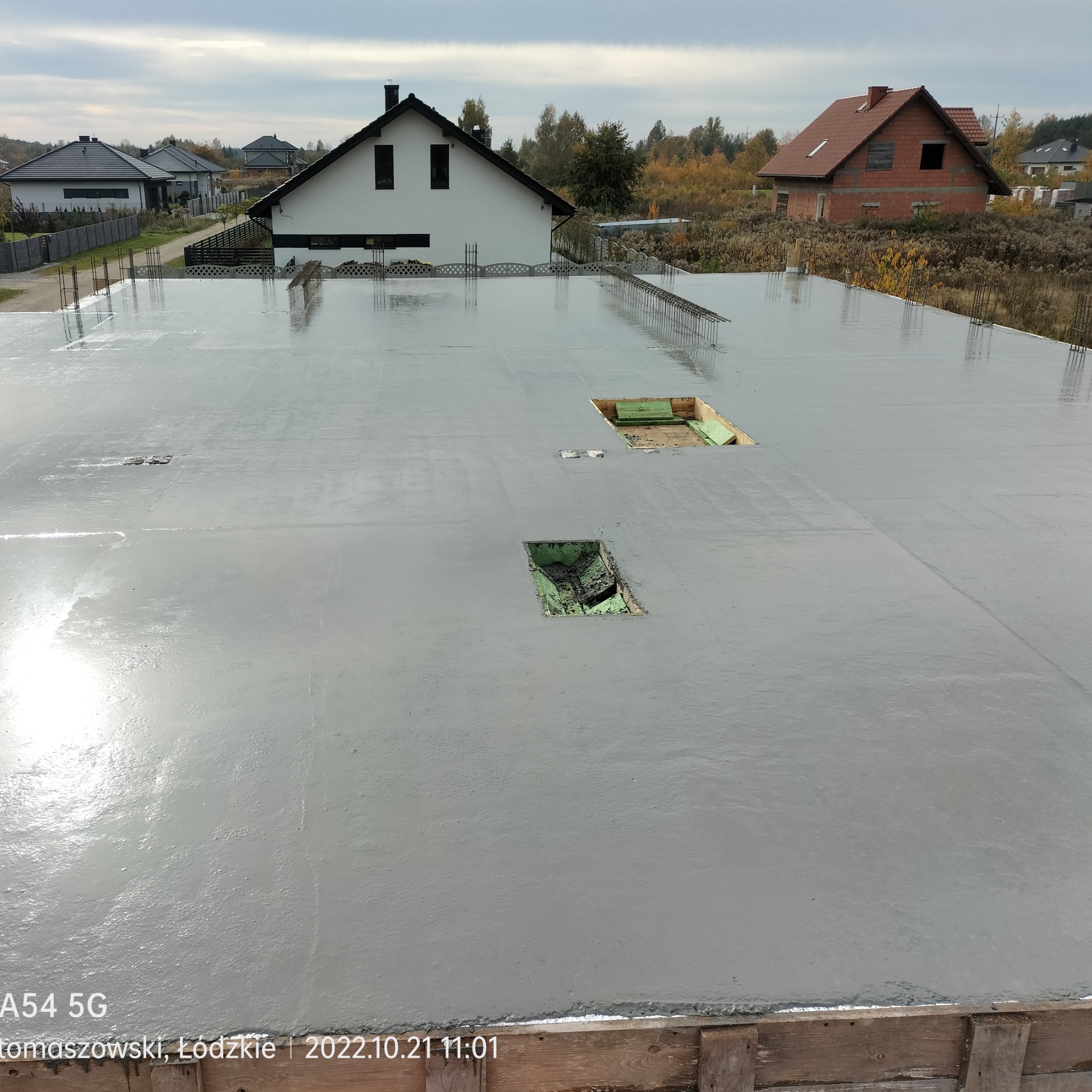 Wylany, świeży beton na płycie fundamentowej z widocznymi otworami na instalacje, zbrojenie wystaje w tle, widok na budynki w oddali przy pochmurnej pogodzie.