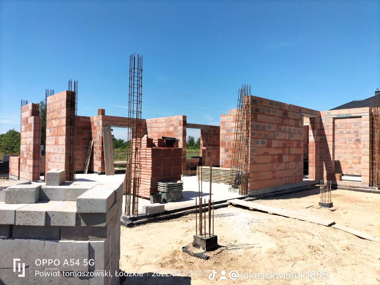 Wznoszenie ścian domu z czerwonej cegły, widoczne zbrojenia pionowe i elementy szalunków, teren budowy w słoneczny dzień.