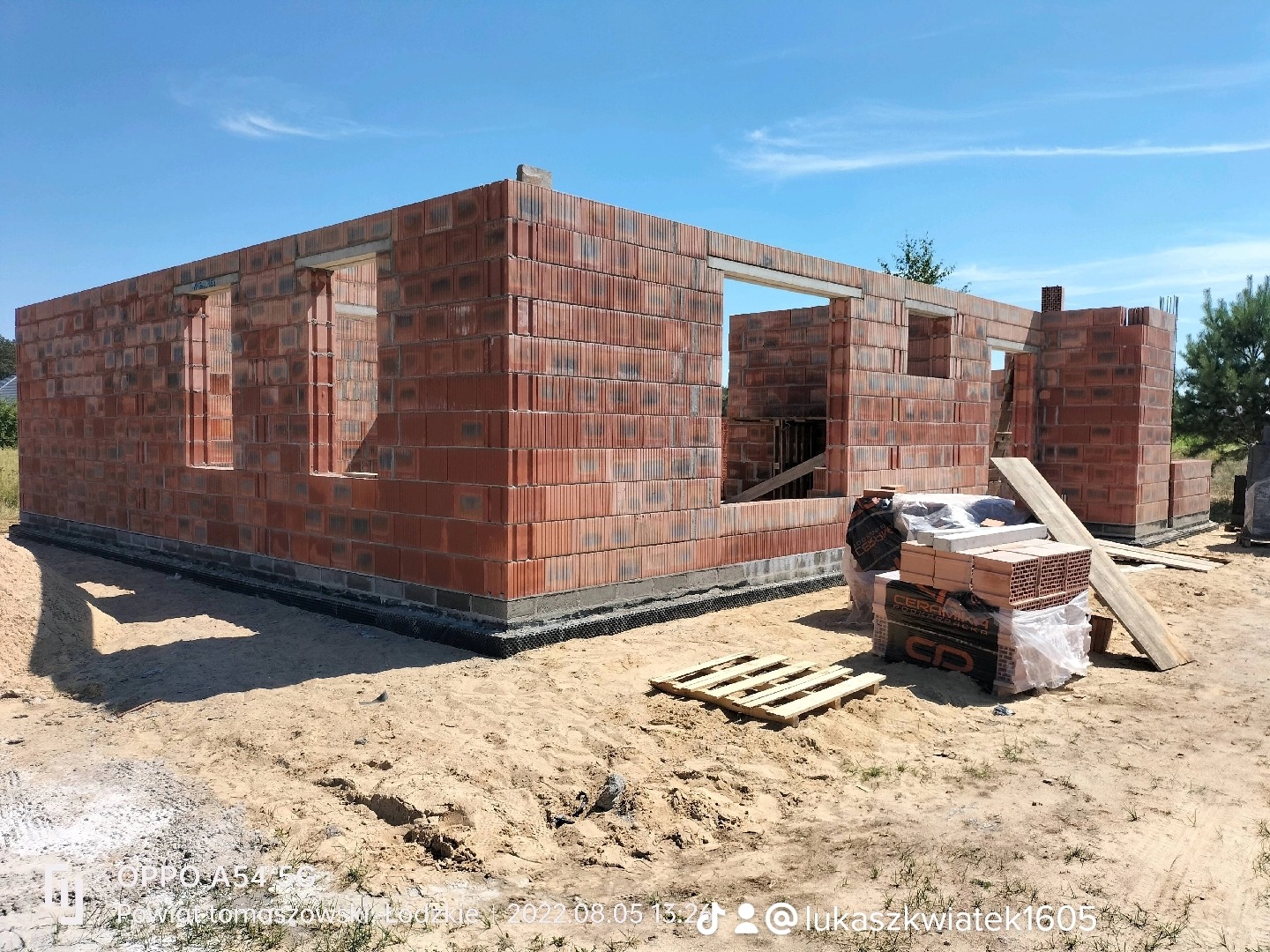 Wznoszenie ścian zewnętrznych domu jednorodzinnego z czerwonej cegły ceramicznej, widoczne otwory okienne i drzwiowe, paleta z cegłami na pierwszym planie, teren budowy.