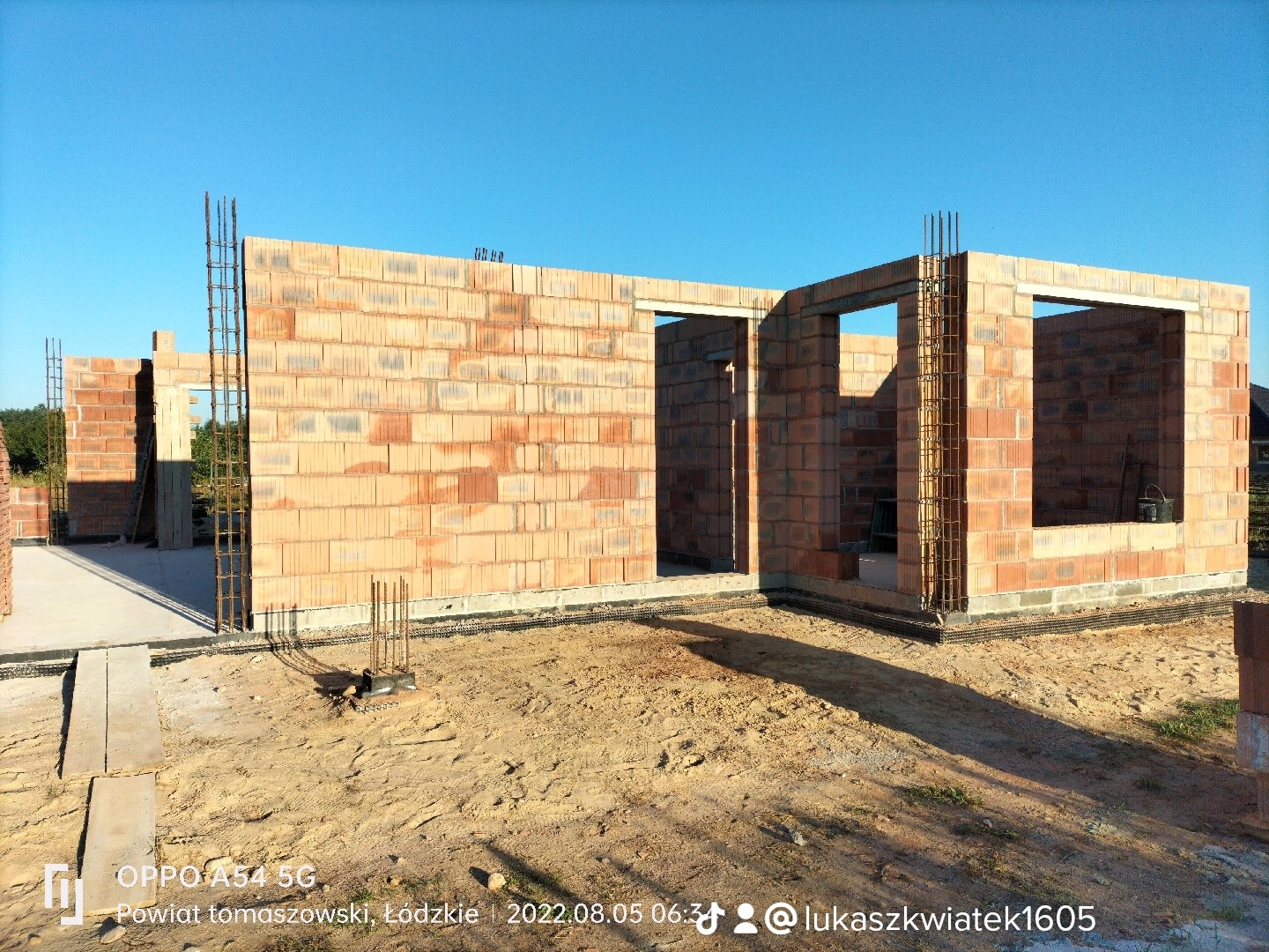 Wznoszenie ścian parterowego budynku z czerwonej cegły ceramicznej, widoczne zbrojenia w narożnikach, przygotowane otwory okienne i drzwiowe, teren budowy w słoneczny dzień.