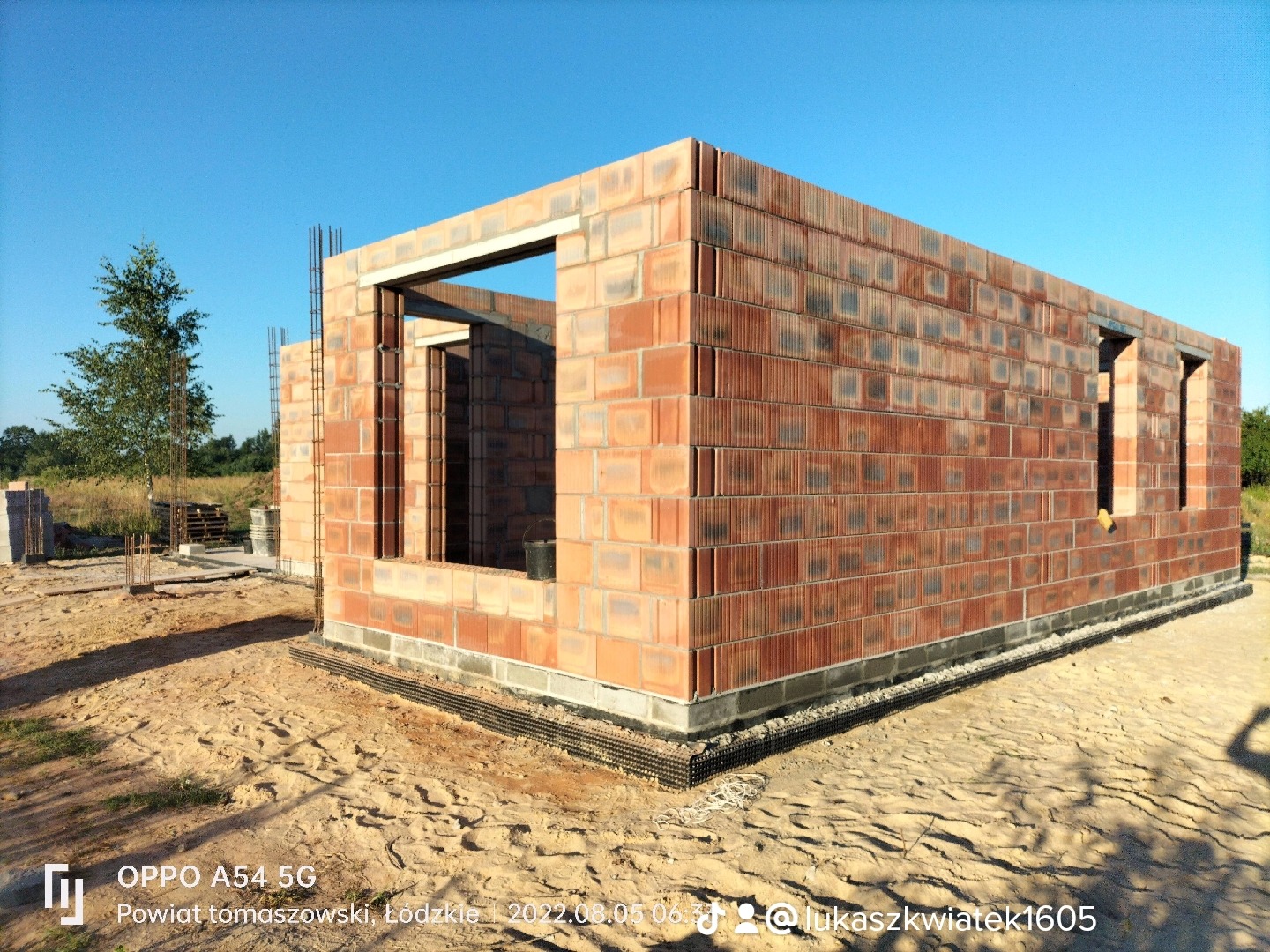 Wznoszenie ścian zewnętrznych z czerwonej cegły modularnej na fundamencie z izolacją, widoczne otwory okienne i drzwiowe, zbrojenie pionowe w narożach, teren budowy w słoneczny dzień.