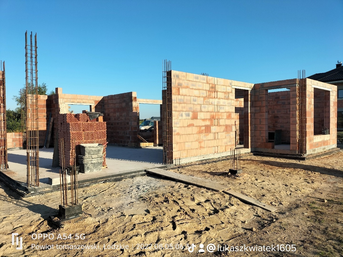 Wznoszenie ścian parterowego domu z czerwonej cegły, widoczne zbrojenia słupów i stos cegieł na fundamencie, w tle błękitne niebo.