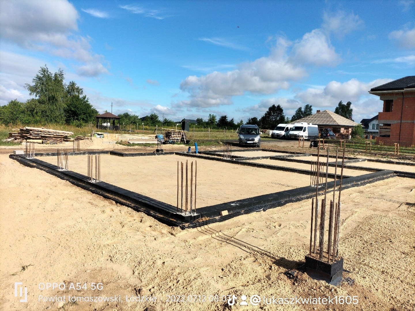 Przygotowany fundament pod dom jednorodzinny z wystającymi prętami zbrojeniowymi, zabezpieczony czarną folią izolacyjną, na piaszczystym podłożu, z widocznymi samochodami dostawczymi w tle.