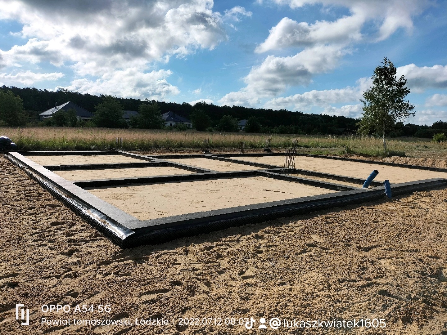 Przygotowany szalunek zbrojeniowy fundamentów na piaszczystym terenie z widocznymi wystającymi prętami zbrojeniowymi i rurami drenażowymi, w tle zabudowa jednorodzinna i drzewa.