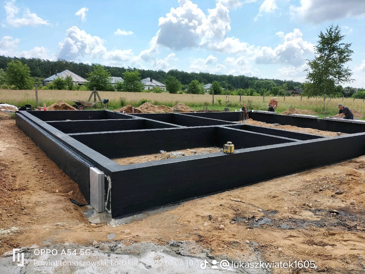 Przygotowany szalunek fundamentów domu jednorodzinnego, pomalowany na czarno, z widoczną izolacją pionową, w tle krajobraz z domami i błękitnym niebem.