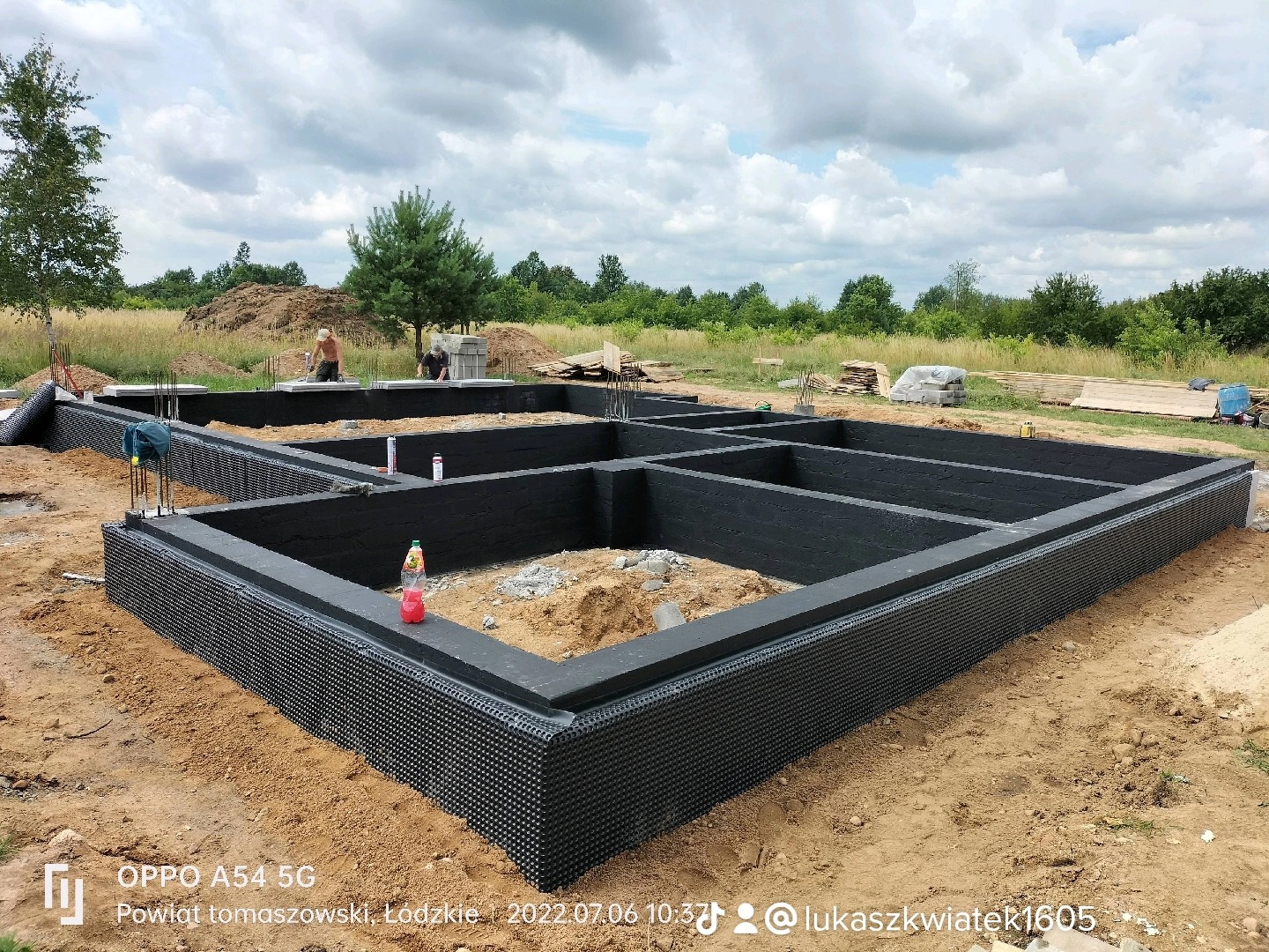 Zabezpieczone fundamenty domu jednorodzinnego z izolacją przeciwwilgociową i termiczną, widoczne zbrojenia, na drugim planie dwóch pracowników budowlanych.