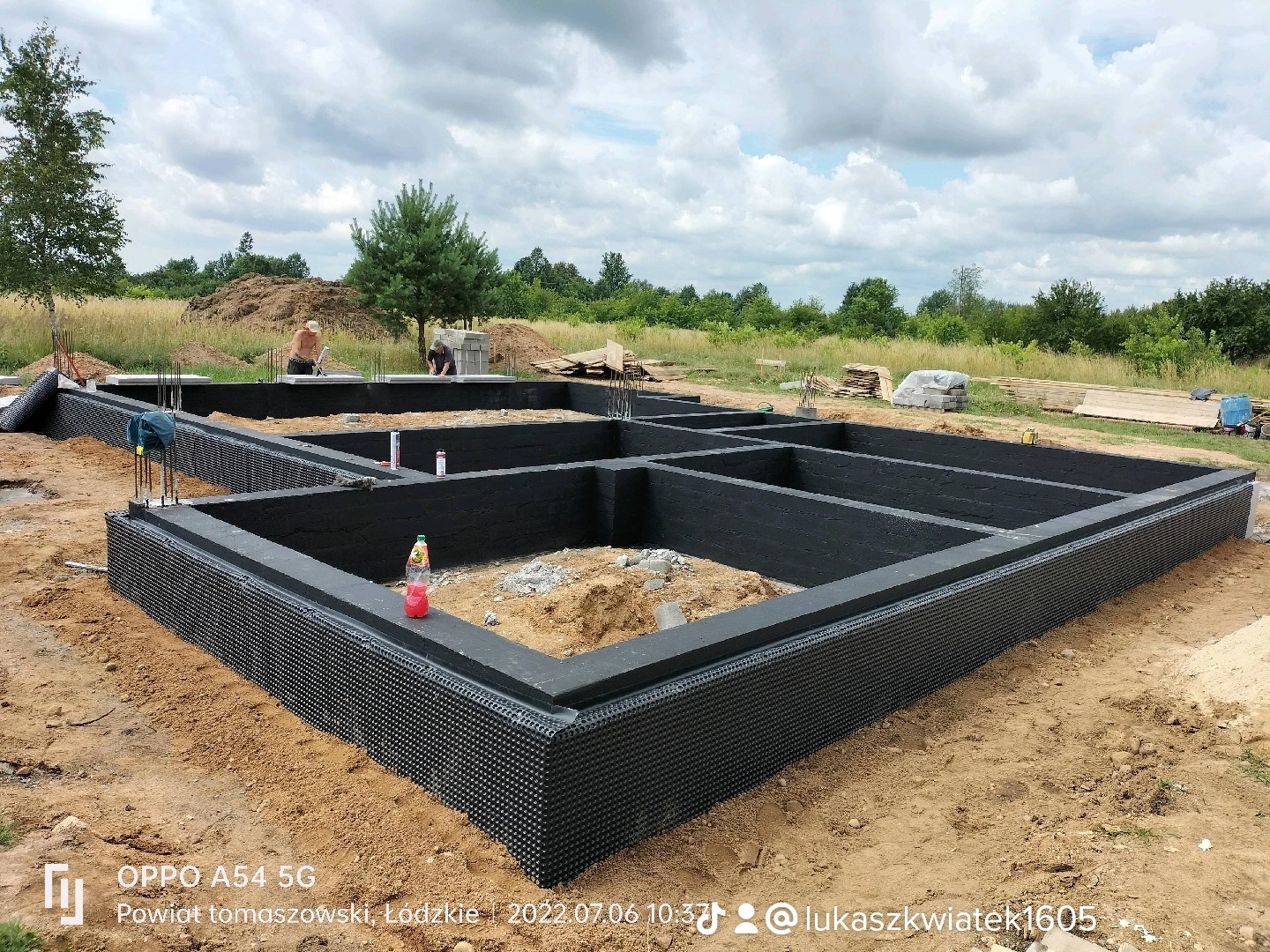 Zabezpieczone przed wilgocią fundamenty domu jednorodzinnego z hydroizolacją i folią kubełkową, widoczne zbrojenia i dwóch pracowników w tle.