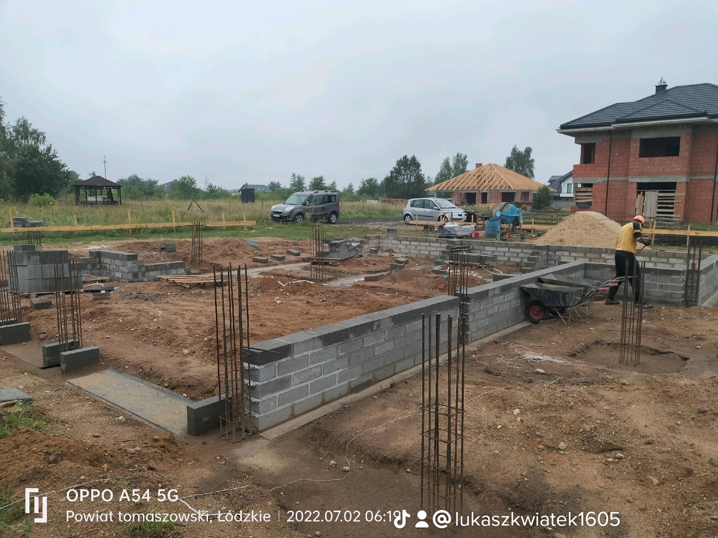 Budowa fundamentów z bloczków betonowych, zbrojenie pionowe wystające z ziemi, taczka i pracownik w kasku na budowie, w tle dom w stanie surowym otwartym i krajobraz podmiejski.