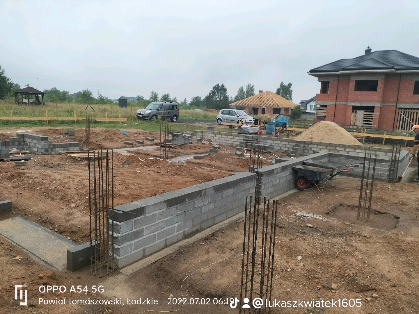 Budowa domu jednorodzinnego w trakcie realizacji: wylane fundamenty z wystającymi prętami zbrojeniowymi, częściowo wymurowane ściany fundamentowe z bloczków betonowych, na działce piasek, taczka,...