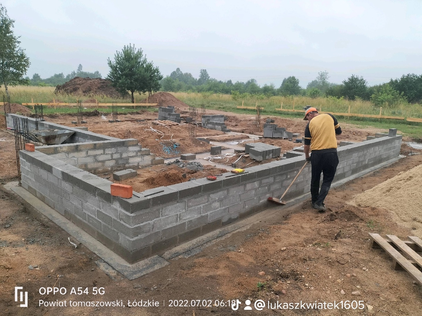 Fundamenty domu z bloczków betonowych, pracownik z miotłą czyści teren wokół, widoczne zbrojenia i wykop pod dalszą część fundamentów, w tle drzewa i składowana ziemia.