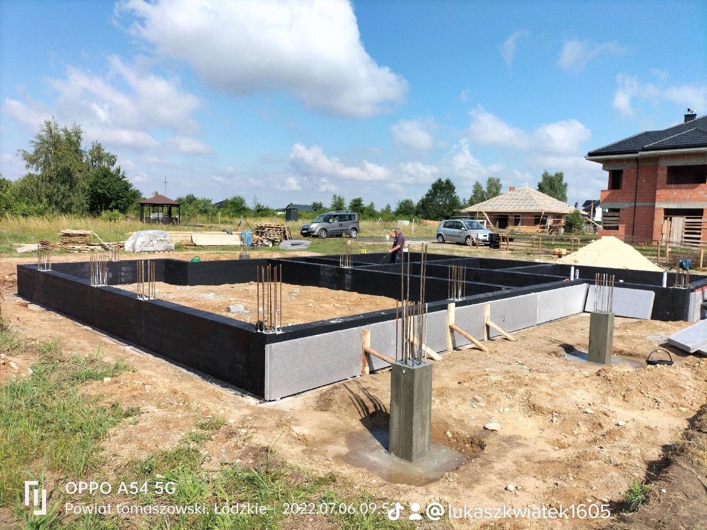 Zalane betonem fundamenty domu jednorodzinnego z wystającymi prętami zbrojeniowymi i zaizolowanymi ścianami zewnętrznymi, widok na budowę w słoneczny dzień.