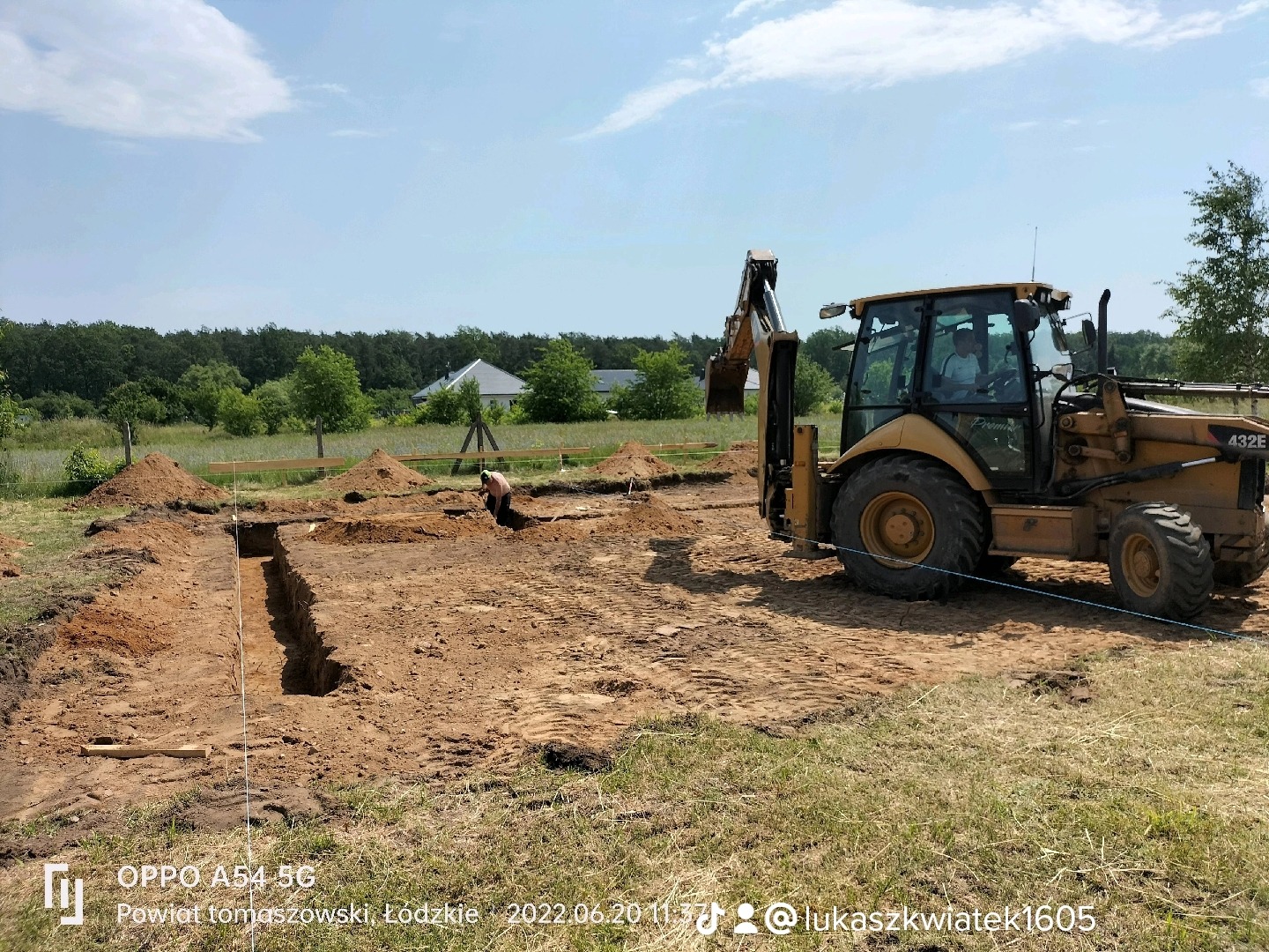 Żółta koparko-ładowarka Case 432E obok wykopu pod fundamenty domu jednorodzinnego, widoczny pracownik w wykopie, w tle dom i drzewa.