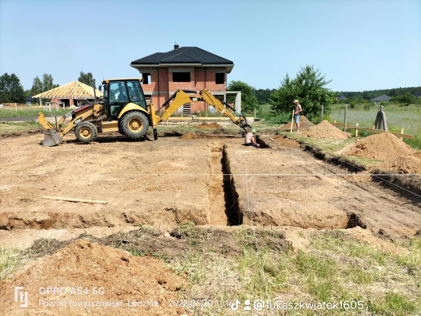Żółta koparko-ładowarka Caterpillar przy wykopie pod fundamenty domu z czerwonej cegły, dwóch pracowników w wykopie, hałdy ziemi, widoczne drewniane szalunki.