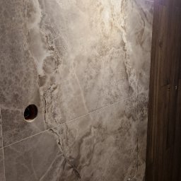 Gre-Fliz - Ściana wyłożona płytkami imitującymi marmur w odcieniach szarości i brązu, widoczne otwory na instalacje elektryczne i wodne, tymczasowe oświetlenie robocze.