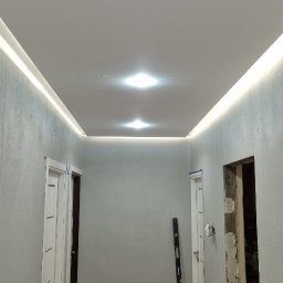 Gre-Fliz - Korytarz w trakcie remontu: wygładzone ściany o szarej fakturze, biały sufit napinany z oświetleniem LED, poziomica oparta o ścianę, widoczne ślady prac na podłodze.