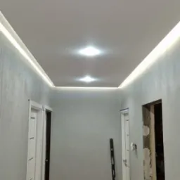 Korytarz w trakcie remontu: wygładzone ściany o szarej fakturze, biały sufit napinany z oświetleniem LED, poziomica oparta o ścianę, widoczne ślady prac na podłodze.