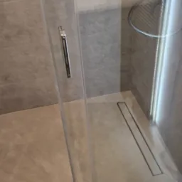 Szklana kabina prysznicowa z odpływem liniowym w nowoczesnej łazience wyłożonej płytkami w odcieniach beżu i szarości, widoczny fragment ściany z dekoracyjną fakturą.