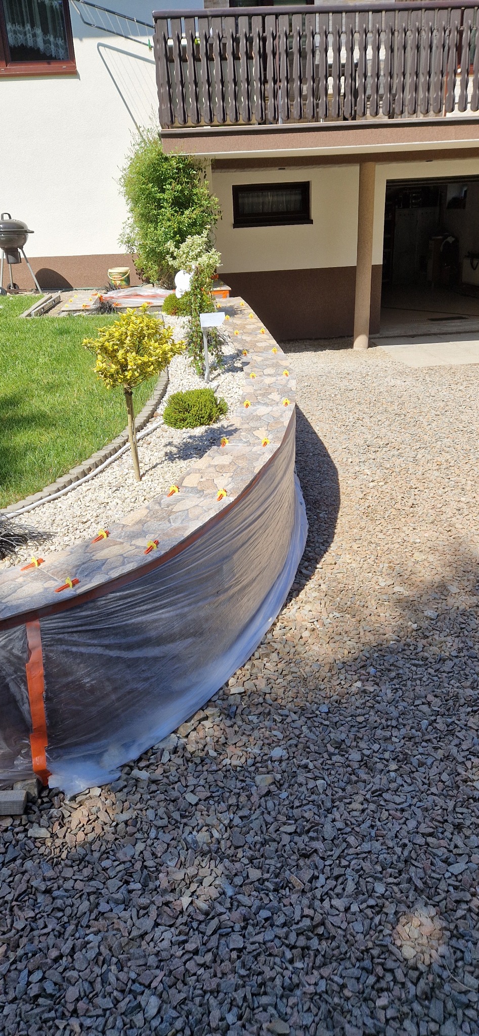 Zakrzywiony murek wykończony płytkami imitującymi kamień, zabezpieczony folią ochronną podczas prac wykończeniowych, z widocznymi żółtymi klinami do poziomowania płytek.