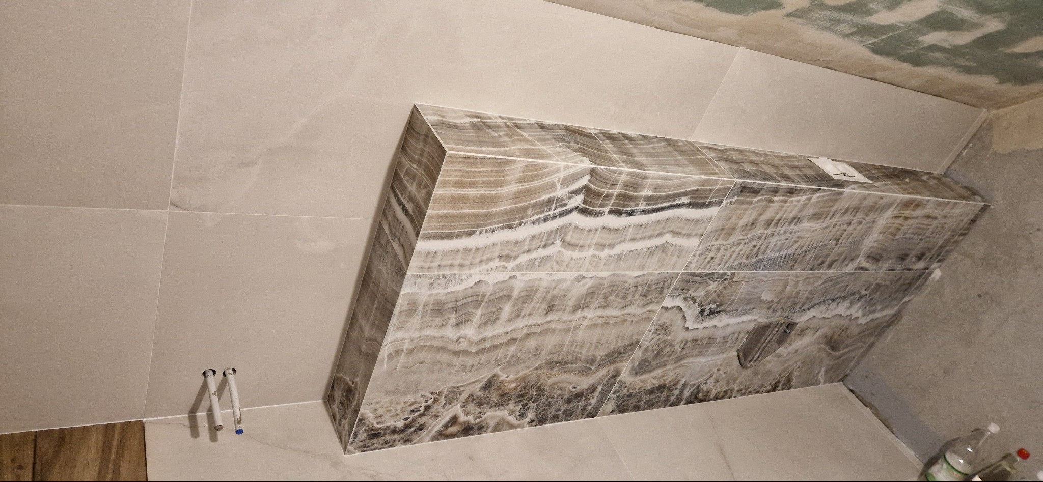 Wykończona wnęka z płytek imitujących onyks w łazience, widoczne rury instalacji wodnej i niedokończony sufit.