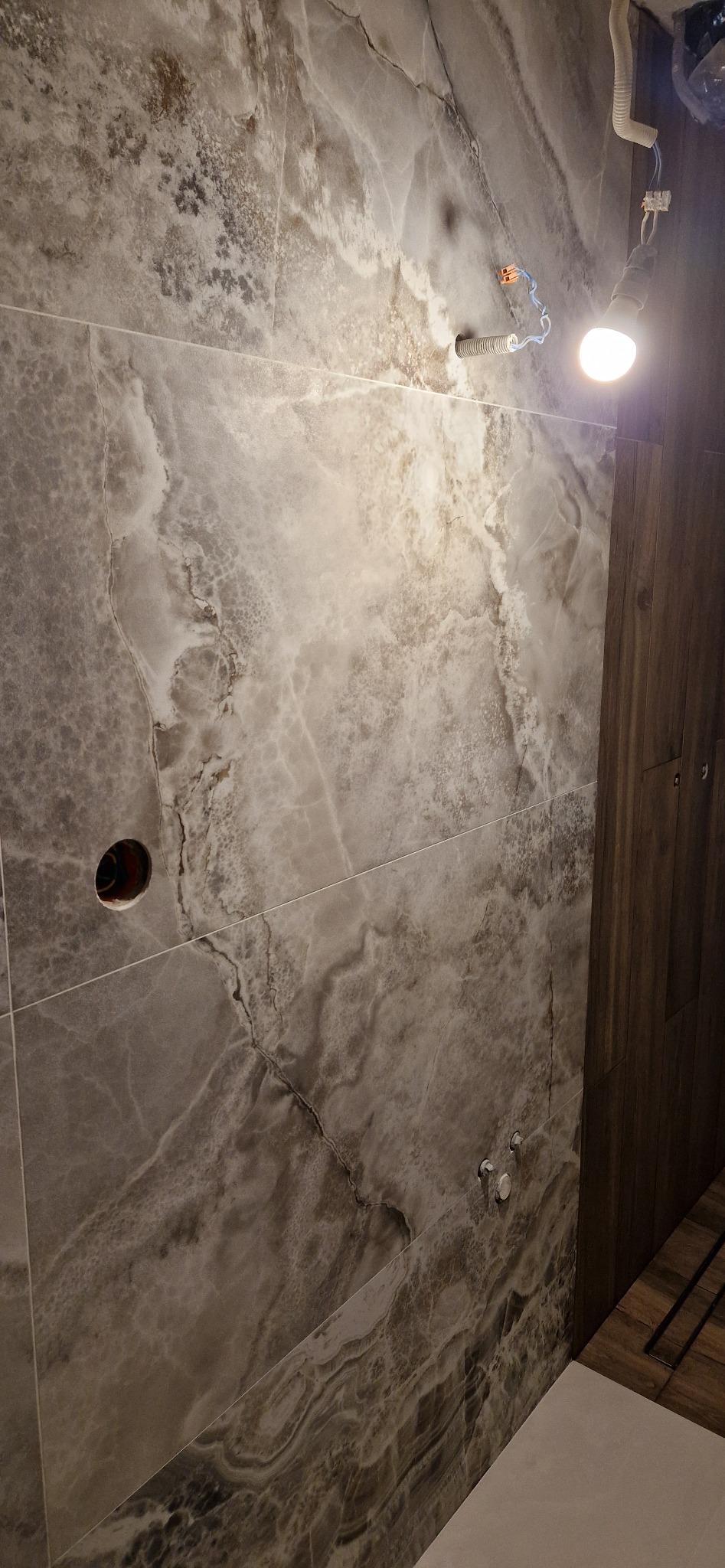 Ściana wyłożona płytkami imitującymi marmur w odcieniach szarości i brązu, widoczne otwory na instalacje elektryczne i wodne, tymczasowe oświetlenie robocze.