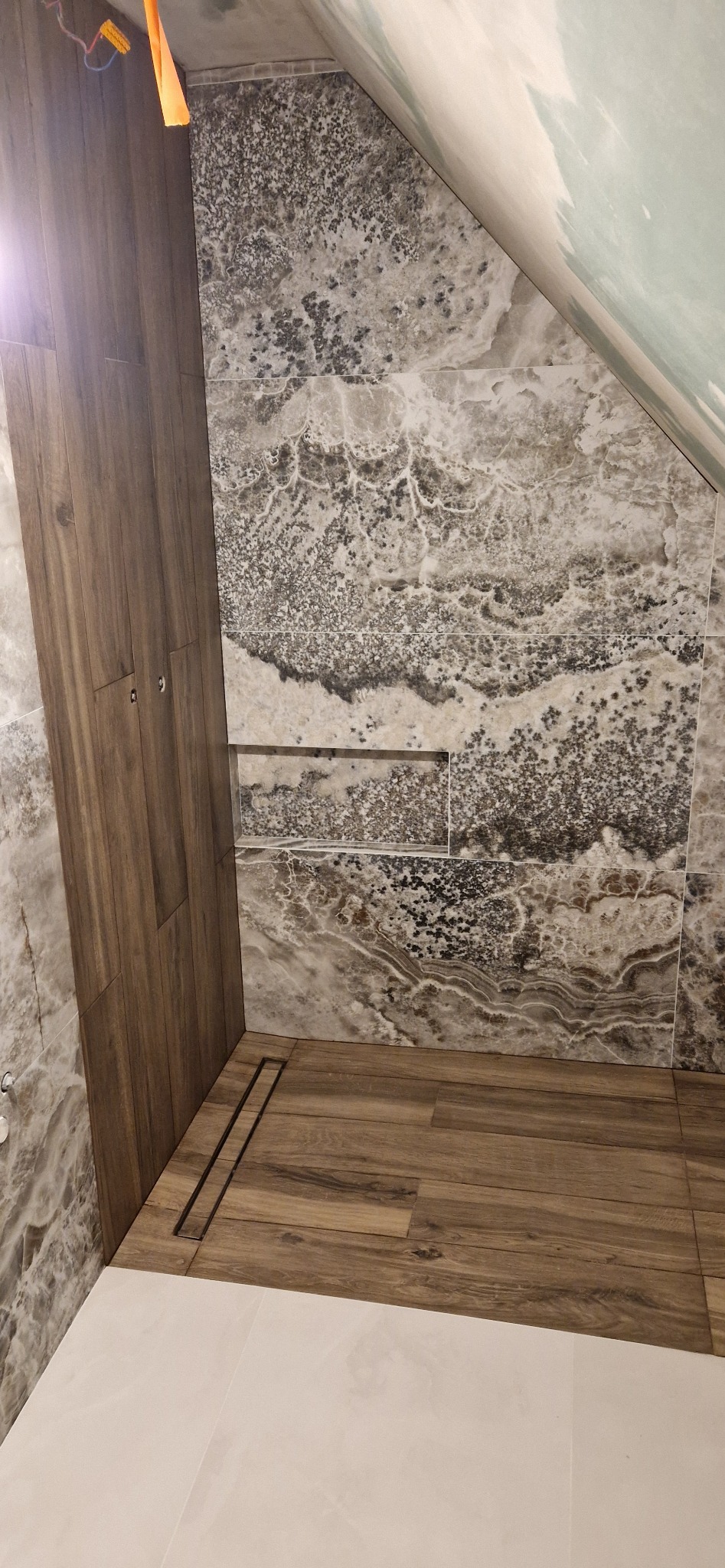 Wykończona kabina prysznicowa z odpływem liniowym, drewnopodobną podłogą i ścianami z płytek imitujących marmur w odcieniach szarości i beżu, wnęka w ścianie wykończona tym samym materiałem.