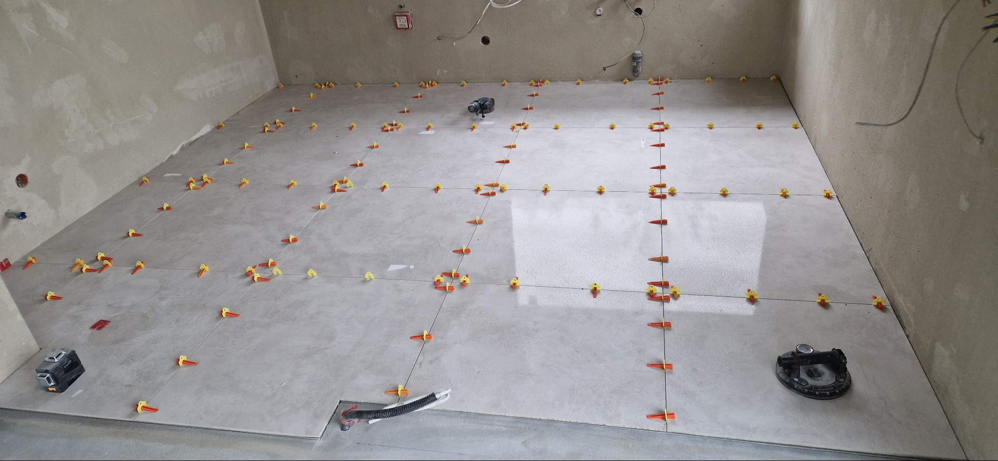 Układanie jasnych płytek podłogowych w surowym wnętrzu, widoczne poziomice laserowe i system poziomowania płytek z żółtymi klinami.