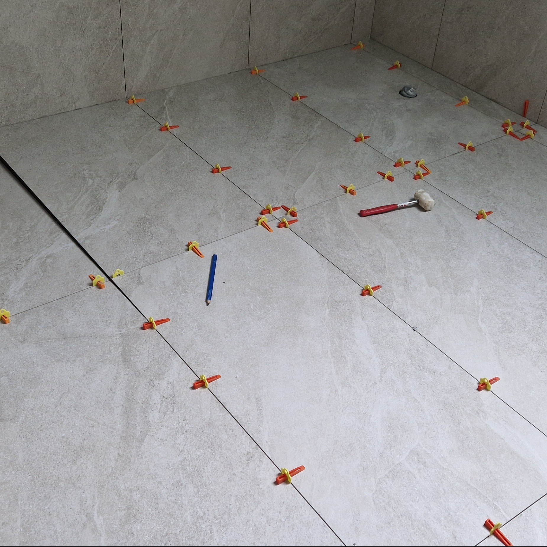 Układanie płytek podłogowych w łazience z użyciem systemu poziomowania: pomarańczowe klipsy i czarne sznurki wyznaczają linie, obok leży gumowy młotek i niebieski ołówek.