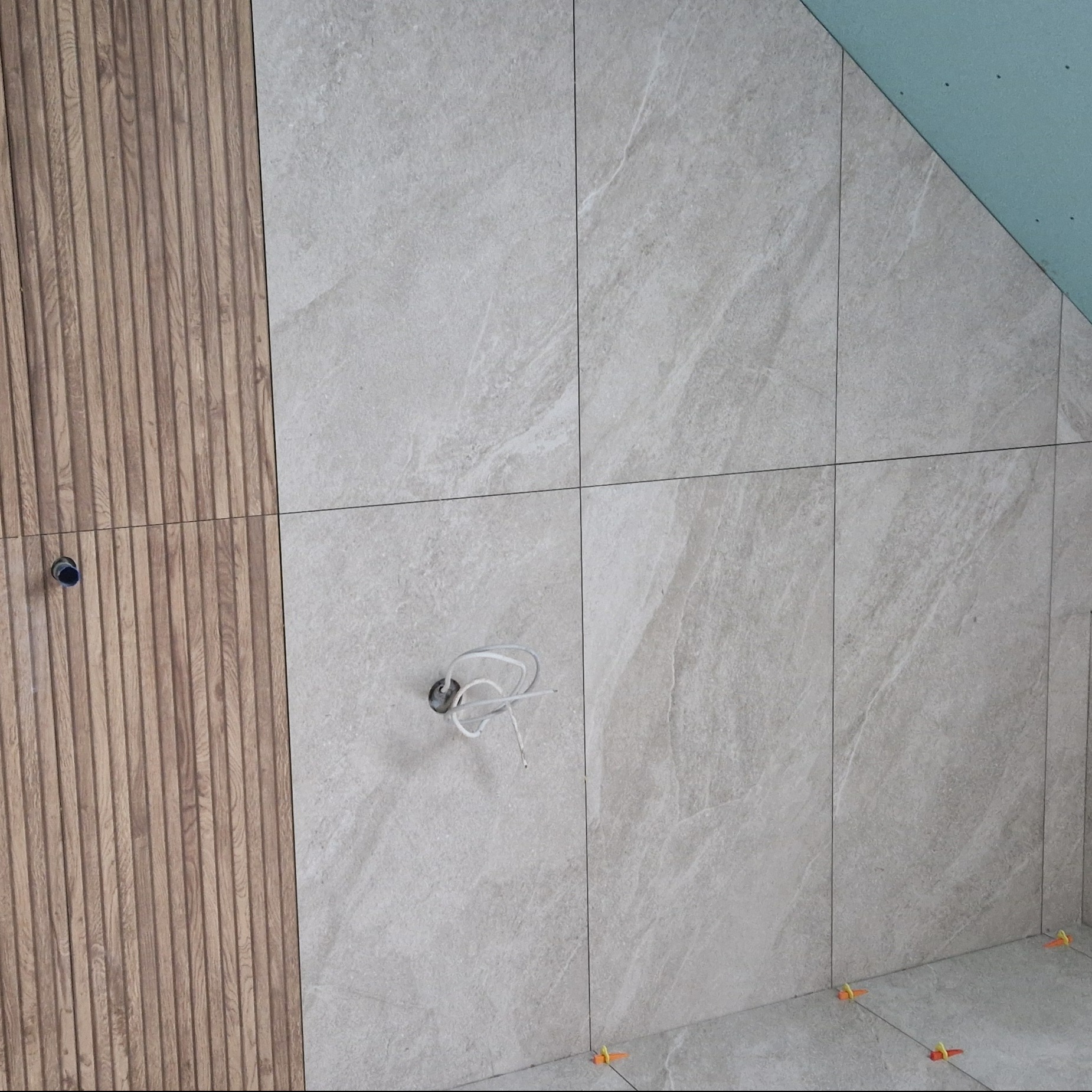 Ściana wyłożona płytkami imitującymi kamień i drewnianymi panelami ściennymi; widoczne przewody elektryczne i poziomujące klipsy do płytek.