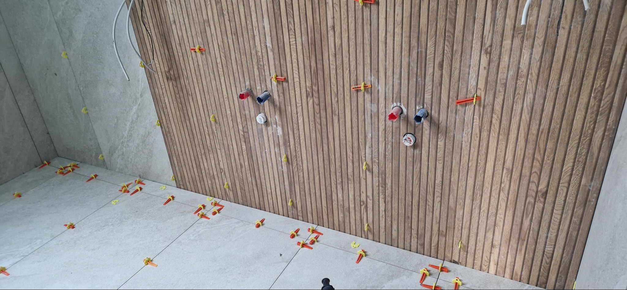 Montaż płytek na podłodze i ścianie z drewnopodobnymi panelami w łazience, widoczne krzyżyki dystansowe i otwory pod armaturę.