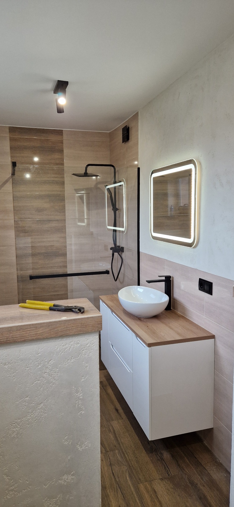 Nowoczesna łazienka z kabiną prysznicową typu walk-in, umywalką nablatową na białej szafce z blatem imitującym drewno oraz podświetlanym lustrem; na blacie obok kabiny leżą żółte klucze hydrauliczne.