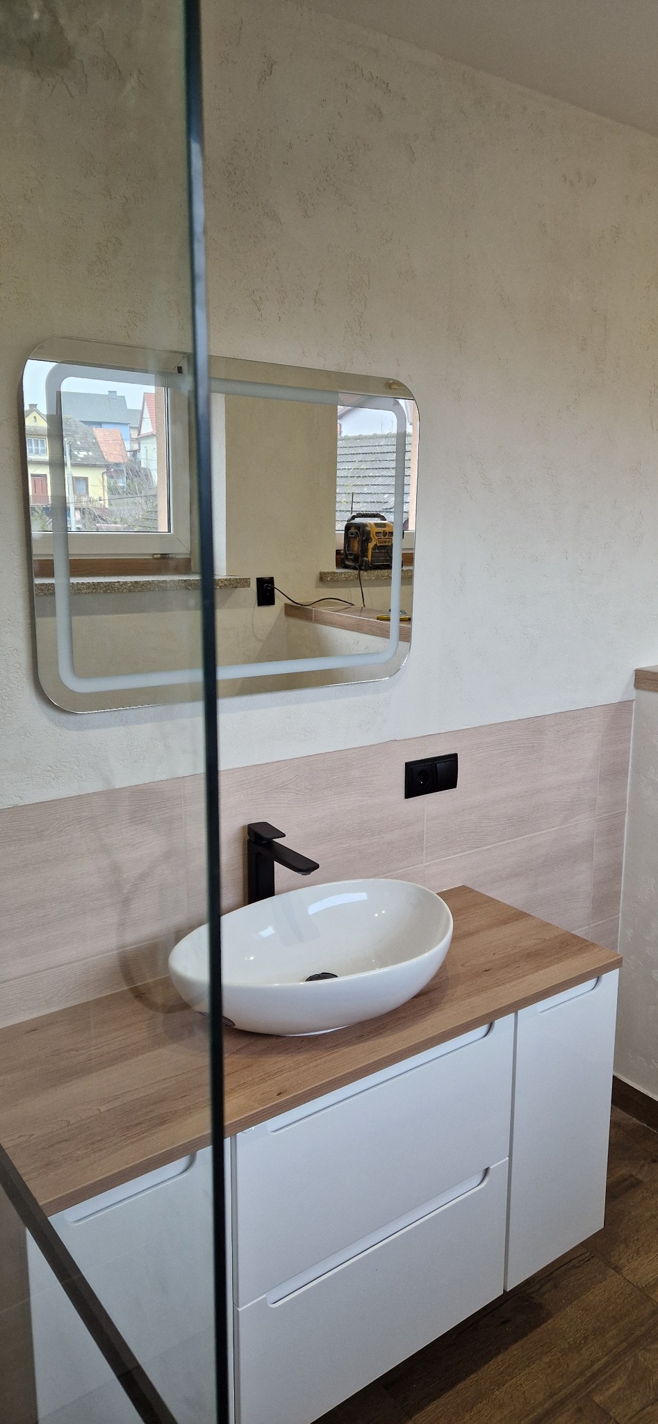 Nowoczesna łazienka z owalną umywalką nablatową, czarną armaturą i podświetlanym lustrem, widok przez szklaną ściankę kabiny prysznicowej, z narzędziem (poziomicą) widocznym w odbiciu lustra.