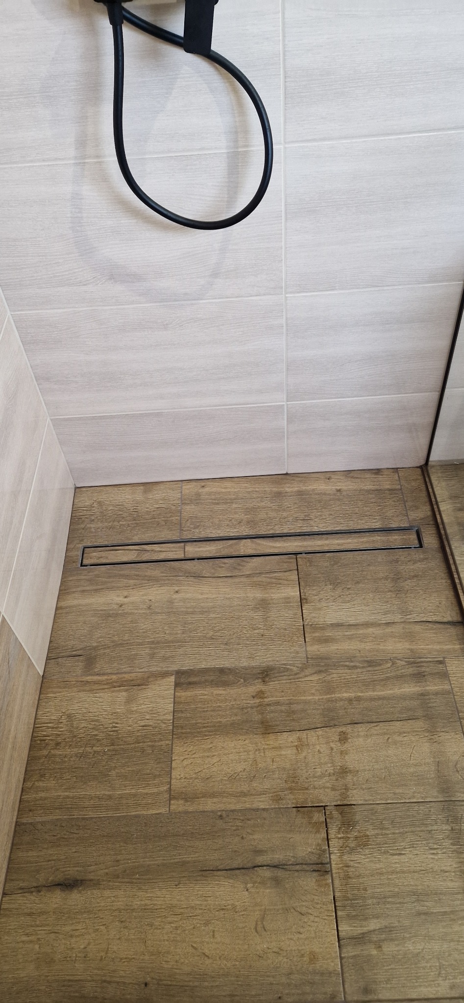 Nowoczesna kabina prysznicowa z odpływem liniowym i płytkami imitującymi drewno na podłodze, oraz jasnymi płytkami na ścianach.