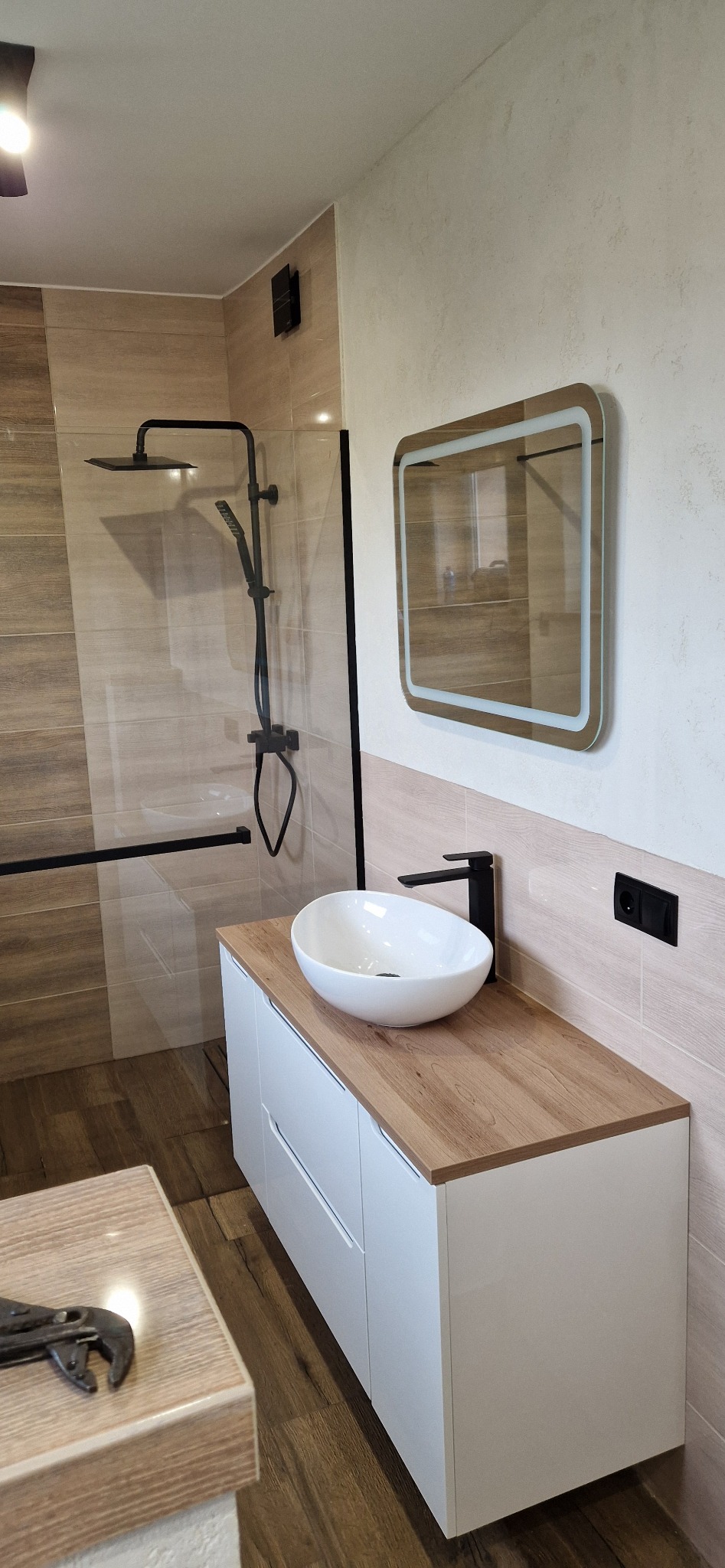 Nowoczesna łazienka z drewnopodobnymi płytkami, kabiną prysznicową typu walk-in z czarną armaturą, umywalką nablatową na szafce z drewnianym blatem i podświetlanym lustrem, na pierwszym planie...