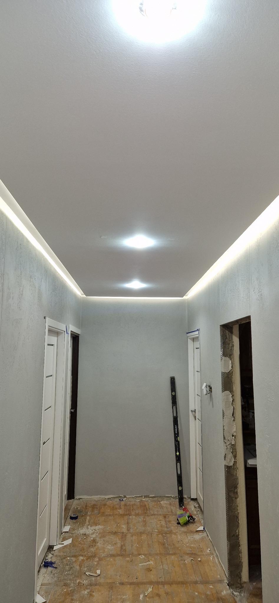 Korytarz w trakcie remontu: wygładzone ściany o szarej fakturze, biały sufit napinany z oświetleniem LED, poziomica oparta o ścianę, widoczne ślady prac na podłodze.