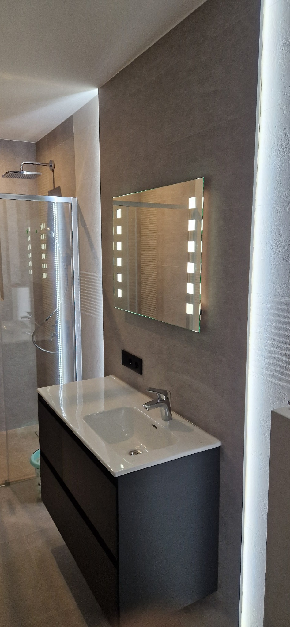 Nowoczesna łazienka z szarą płytką, podświetlanym lustrem i umywalką z szafką w kolorze antracytowym, widoczna kabina prysznicowa z deszczownicą.