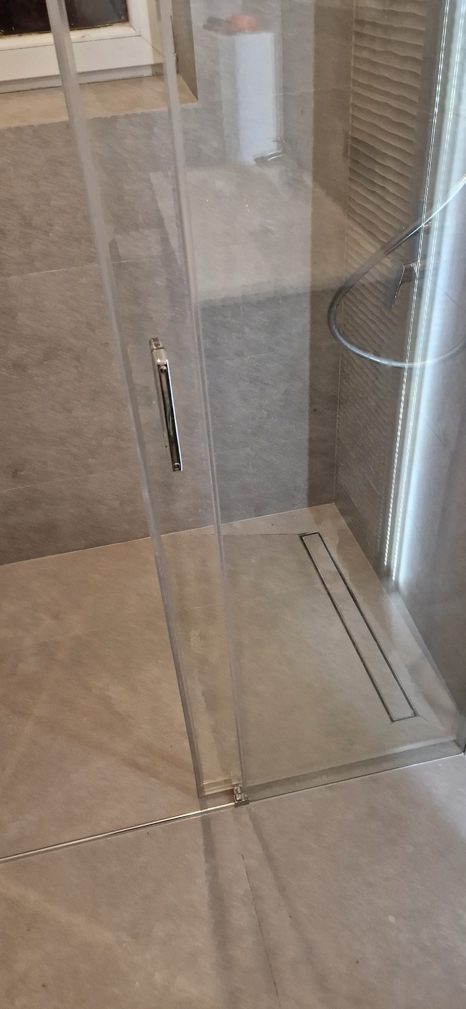 Szklana kabina prysznicowa z odpływem liniowym w nowoczesnej łazience wyłożonej płytkami w odcieniach beżu i szarości, widoczny fragment ściany z dekoracyjną fakturą.