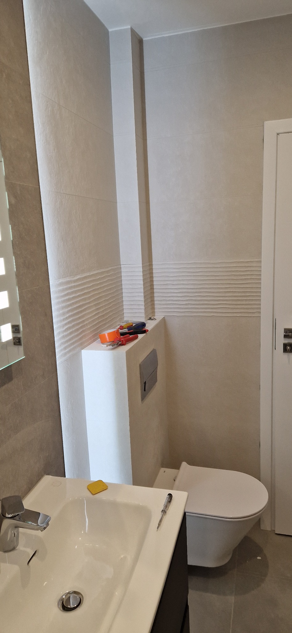 Biała umywalka z baterią i toaleta w jasnej łazience z częściowo ułożonymi płytkami i narzędziami na zabudowie toalety.