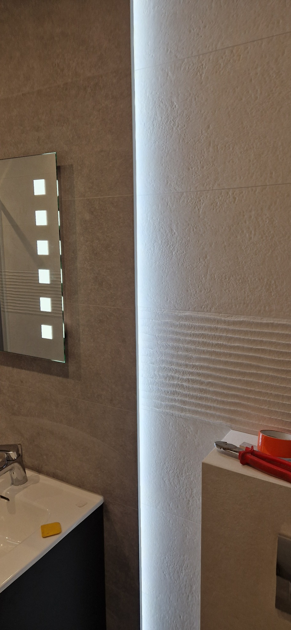 Nowoczesna łazienka z podświetlanym lustrem, beżowymi płytkami o zróżnicowanej fakturze oraz narzędziami na blacie, sugerującymi prace wykończeniowe.