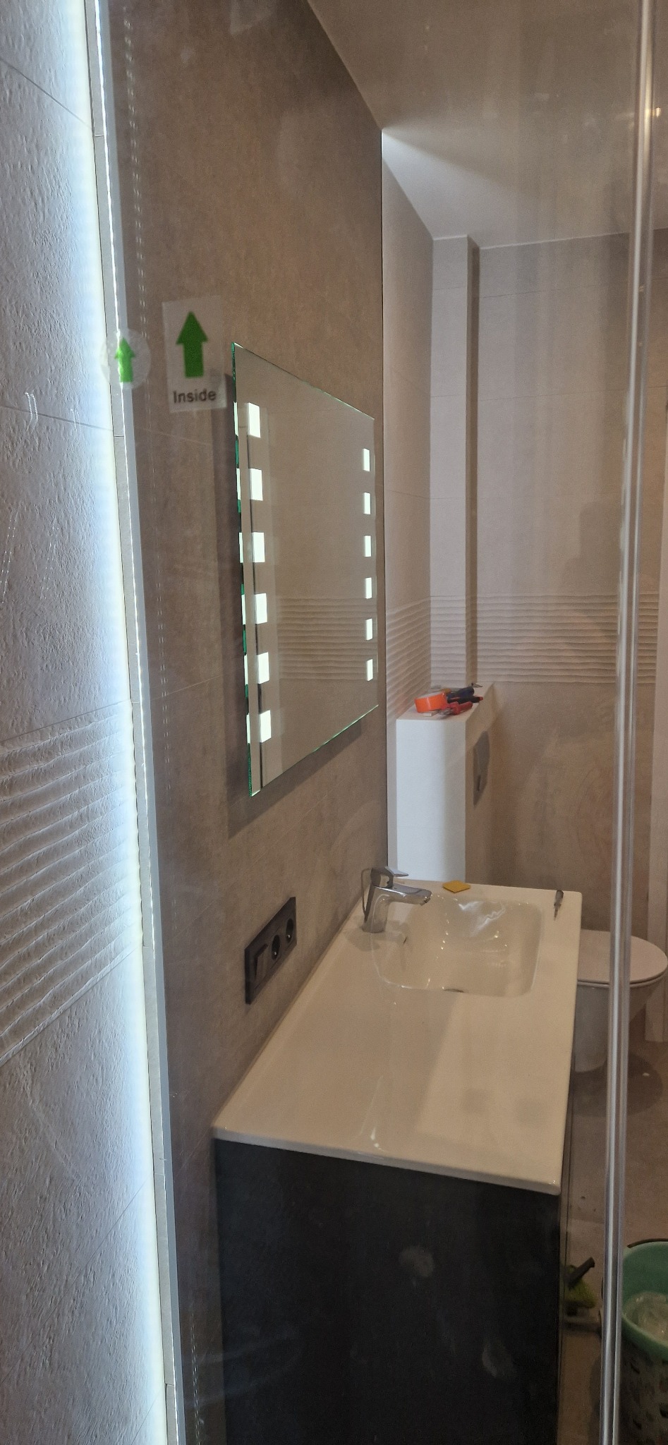 Nowoczesna łazienka z podświetlanym lustrem, umywalką nablatową i szklaną kabiną prysznicową, widoczne narzędzia na szafce nad toaletą.