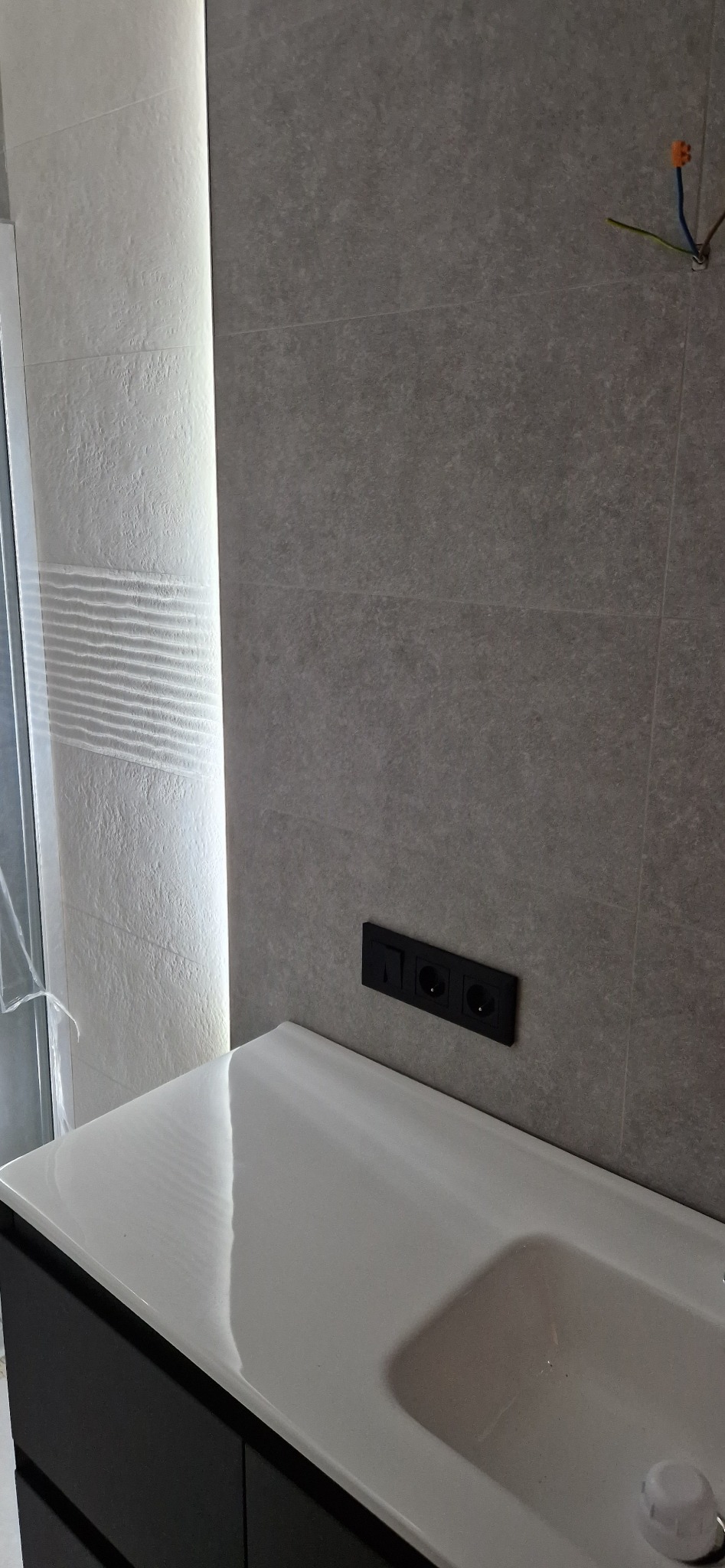 Nowoczesna łazienka w trakcie remontu: szare płytki, biała umywalka z szafką, czarne gniazdka elektryczne, widoczne kable instalacji elektrycznej.