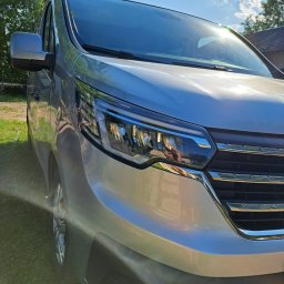 MTM Trans Mariusz Tyszecki - Srebrny bus z nowoczesnymi reflektorami, uchwycony pod kątem z widocznym odbiciem fotografa w lampie, na tle zielonej trawy i błękitnego nieba z promieniami słońca.