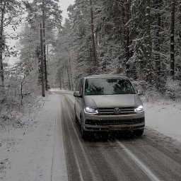 MTM Trans Mariusz Tyszecki - Srebrny bus Volkswagen Transporter jedzie ośnieżoną drogą przez zimowy las, widoczne ślady opon na asfalcie i padający śnieg.