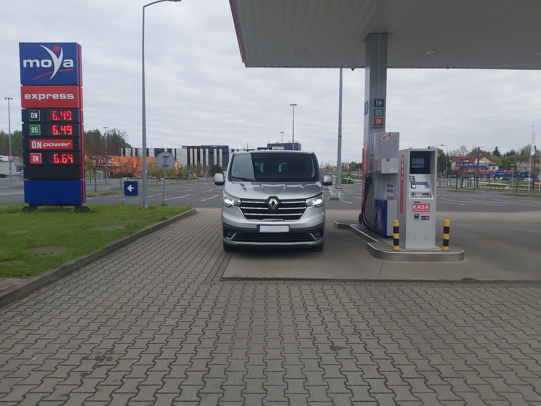 Srebrny van Renault Trafic na stacji paliw Moya, z widocznym cennikiem paliw i dystrybutorem.