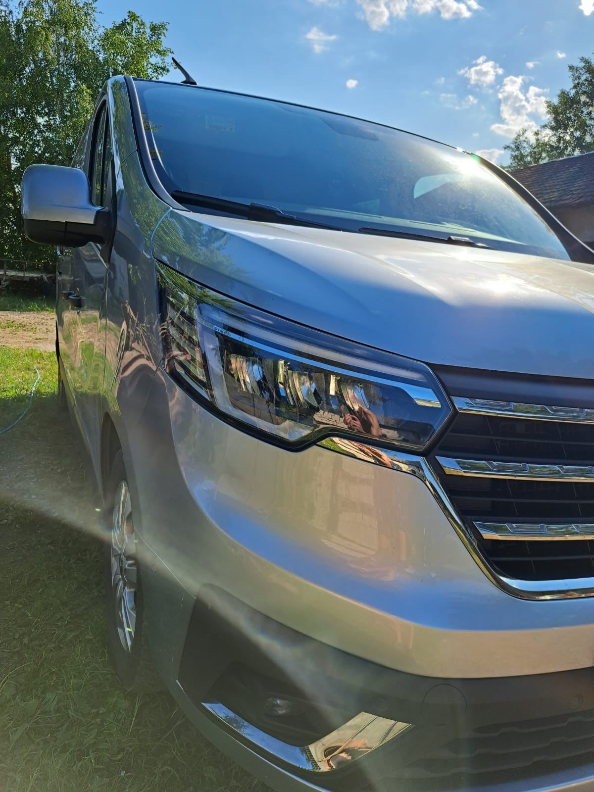 Srebrny bus z nowoczesnymi reflektorami, uchwycony pod kątem z widocznym odbiciem fotografa w lampie, na tle zielonej trawy i błękitnego nieba z promieniami słońca.