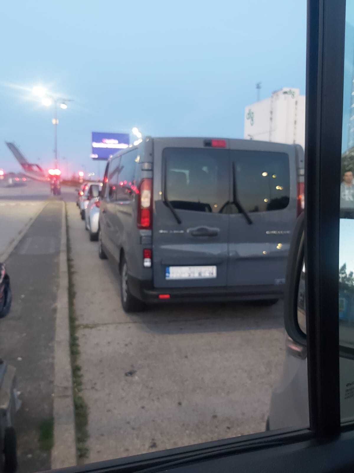 Szary bus zaparkowany na poboczu drogi w pobliżu lotniska, w tle widoczny samolot i budynek lotniskowy.