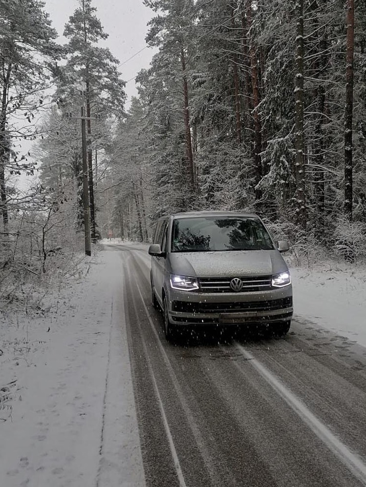 Srebrny bus Volkswagen Transporter jedzie ośnieżoną drogą przez zimowy las, widoczne ślady opon na asfalcie i padający śnieg.