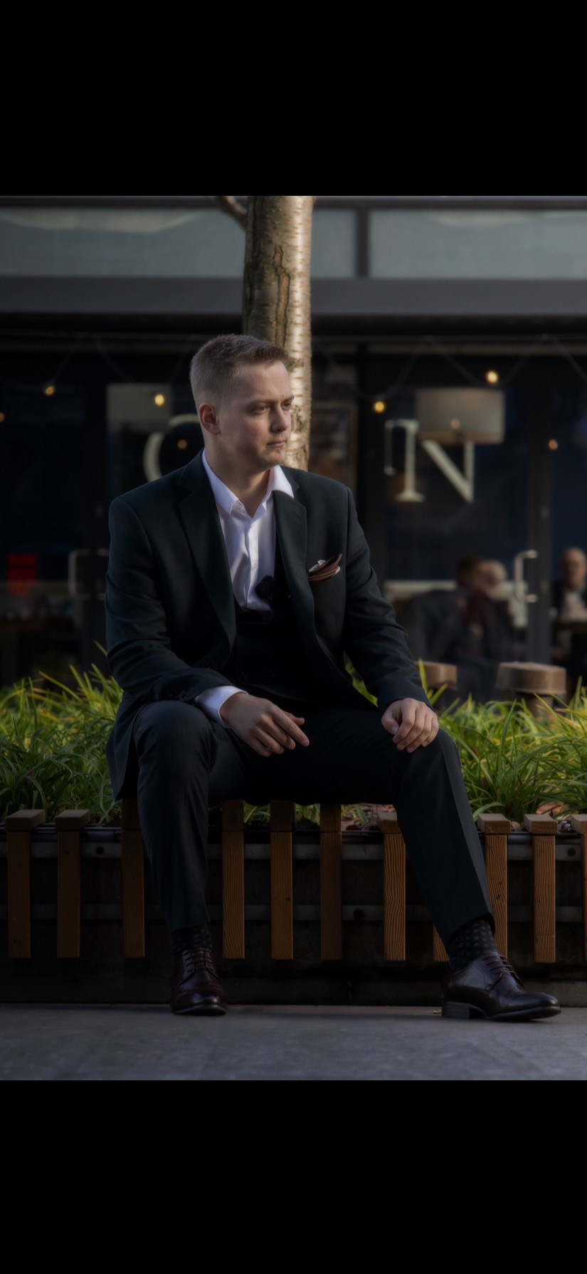 Młody mężczyzna w ciemnym garniturze siedzi na ławce w parku, spoglądając w bok. W tle widoczny budynek z oświetleniem.