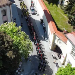 Widok z góry na ulicę z budynkami i drzewami, gdzie maszeruje długa kolumna strażaków i osób z flagami, a na chodnikach stoją obserwatorzy.