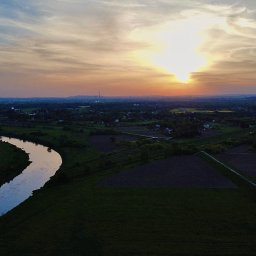 Krystian Nowak - Panoramiczny widok z drona na zakole rzeki, pola uprawne i odległe zabudowania miejskie o zachodzie słońca.
