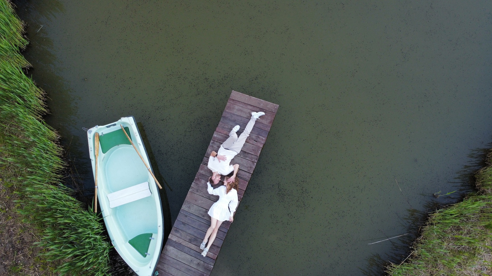 Para w białych ubraniach leży na drewnianym pomoście nad jeziorem, obok łódka z wiosłami, widok z góry.
